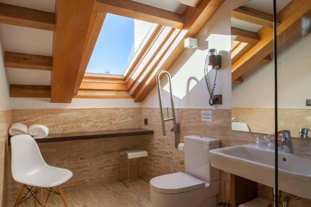 Banheiro moderno do Hotel San Miguel com claraboia, vigas de madeira, vaso sanitário, pia, cadeira, toalhas, secador de cabelo e superfícies de mármore bege. Imagem para ilustrar post sobre hotéis em Santiago de Compostela.