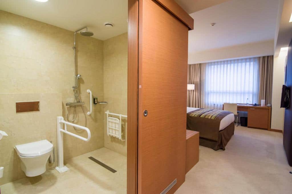 Quarto da Ramada by Wyndham Oradea com banheiro acessível, com barras de apoio, assento no chuveiro e uma cama visível no quarto adjacente.