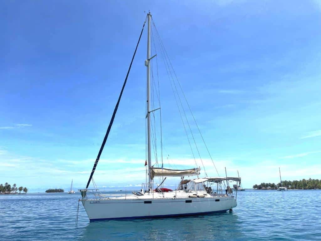 sanblasonsailboats, um veleiro branco flutua em águas azuis calmas sob um céu claro, com ilhas e outros barcos ao fundo.