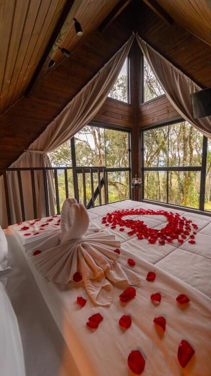 Uma cama nas Cabanas Cold Mountain com pétalas de rosas dispostas em forma de coração e cisnes de toalha dobrada, em um quarto de madeira com janelas grandes e vista para a floresta.