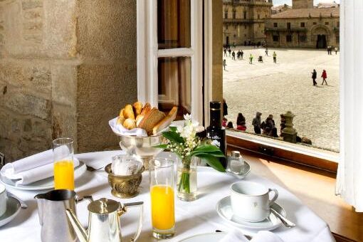 Mesa de café da manhã do Parador de Santiago com bolos e suco de laranja ao lado de uma janela aberta, com vista para uma praça histórica da cidade e para a catedral. Imagem para ilustrar post sobre hotéis em Santiago de Compostela.