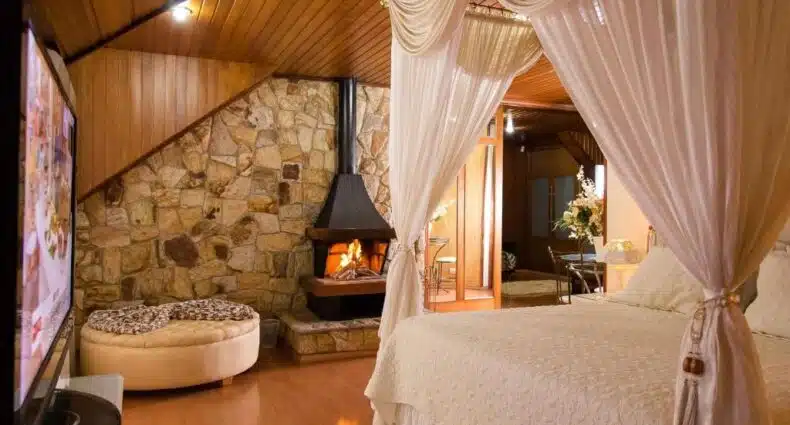 Um quarto com cama de dossel, lareira de pedra, teto com painéis de madeira e uma área de estar com um pufe redondo, da Pousada D'lines com lareira em Campos do Jordão