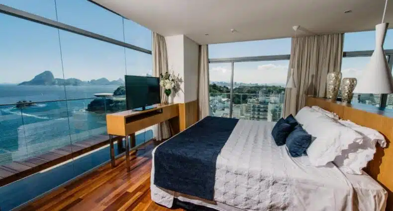Um quarto moderno com janelas do chão ao teto com vista para o oceano e para a paisagem urbana, com uma cama e pisos de madeira, no H Niteroi Hotel.