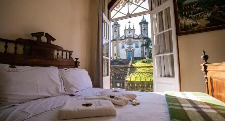 Uma cama feita com toalhas em cima, ao lado de portas abertas que mostram a vista de uma igreja histórica e a vegetação do lado de fora, do Teatro Hotel Boutique. Representa melhores hotéis em Minas Gerais.