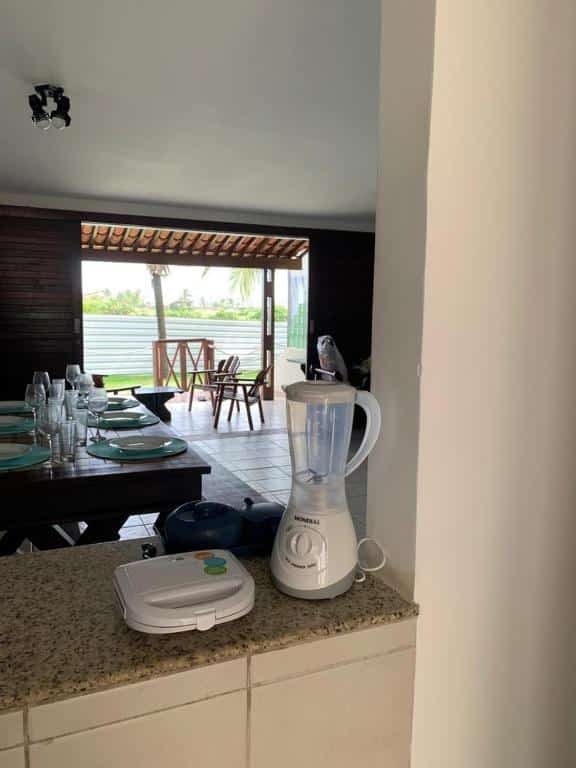 Um liquidificador branco e uma sanduicheira em um balcão de cozinha da Casa no Paraíso dos Carneiros em Tamandaré, com uma mesa de jantar e um pátio visíveis ao fundo.