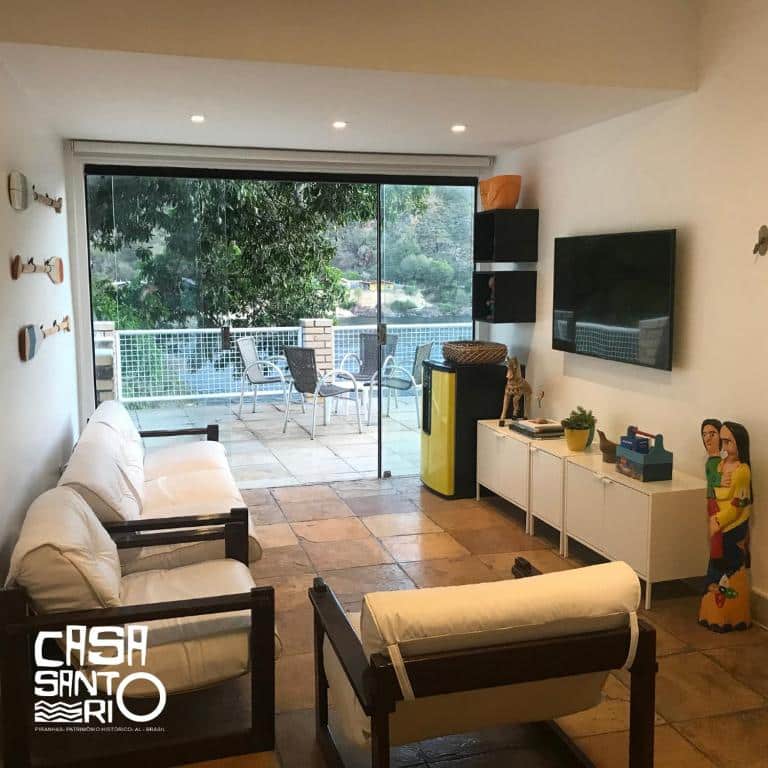 Uma sala de estar com móveis brancos, TV e decoração, que se abre para uma varanda com mesa, cadeiras e vista panorâmica na Casa Santo Rio.