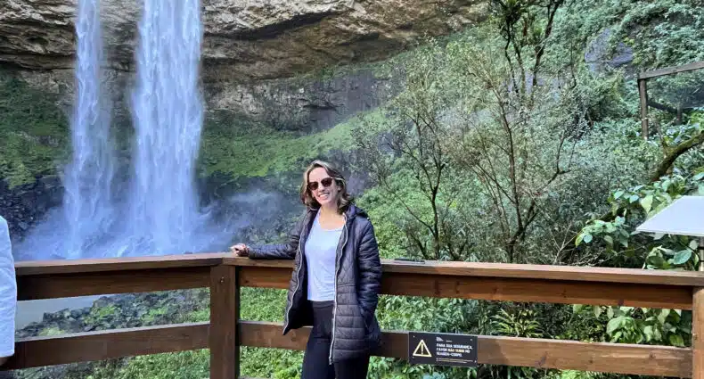 Mulher sorrindo em frente à cachoeira Caracol, em Canela, com jaqueta e calça preta