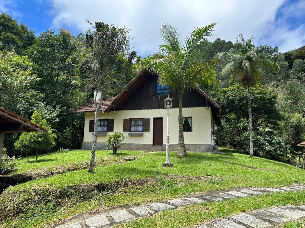 Uma casa de dois andares com telhado marrom, cercada por grama, árvores e uma passarela de pedra sob um céu parcialmente nublado. Foto do chalé chácara caminho do vale