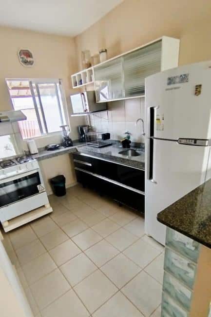 Uma pequena cozinha do Chalet no Paraíso com eletrodomésticos brancos, uma bancada preta, um fogão a gás e uma janela aberta acima da pia.