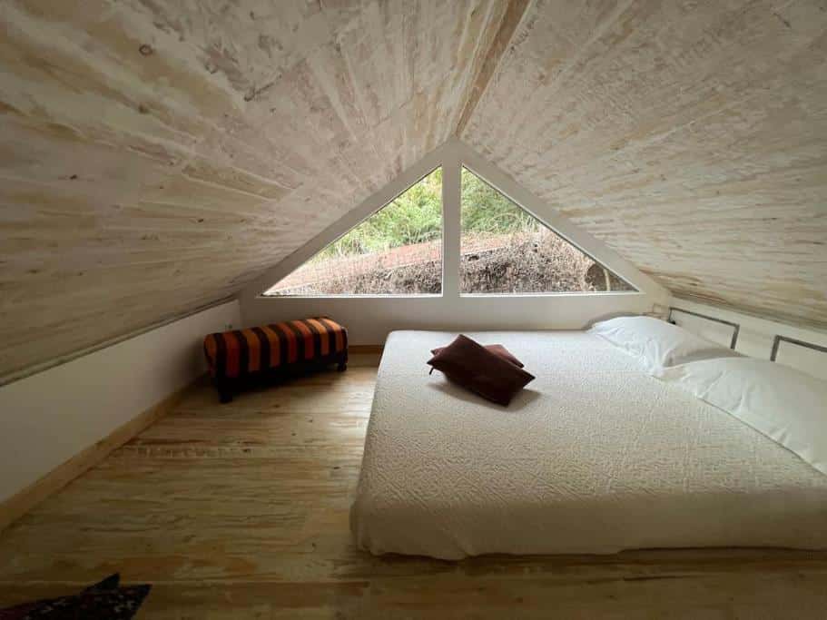 Um pequeno quarto no sótão do Eco-Chic Retreat com uma cama de casal, dois travesseiros, um cobertor e uma janela com vista para a floresta.