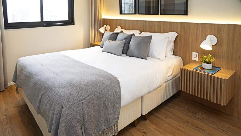 Uma cama bem arrumada no Expand 2958 - Pinheiros com roupa de cama cinza e branca, piso de madeira, mesa de cabeceira e luzes de leitura montadas na parede.