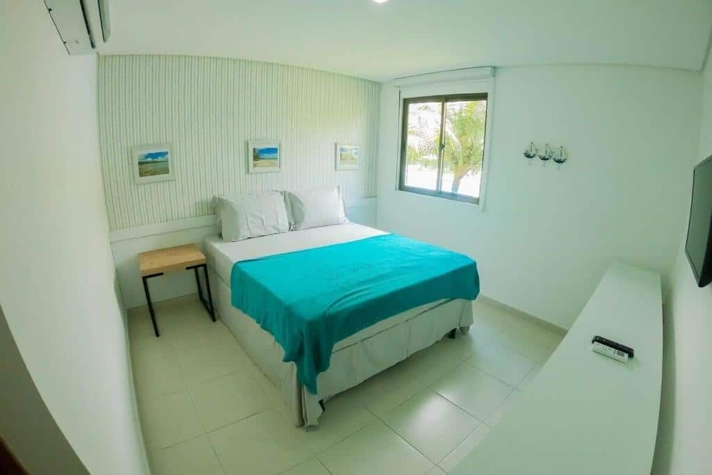 Um quarto pequeno e bem iluminado no Freitas Resort - Carneiros Beach Resort com uma cama de casal, colcha azul-petróleo, janela, arte na parede, TV e um banco de madeira como criado-mudo. Essa é uma das indicações de onde ficar na Praia dos Carneiros.