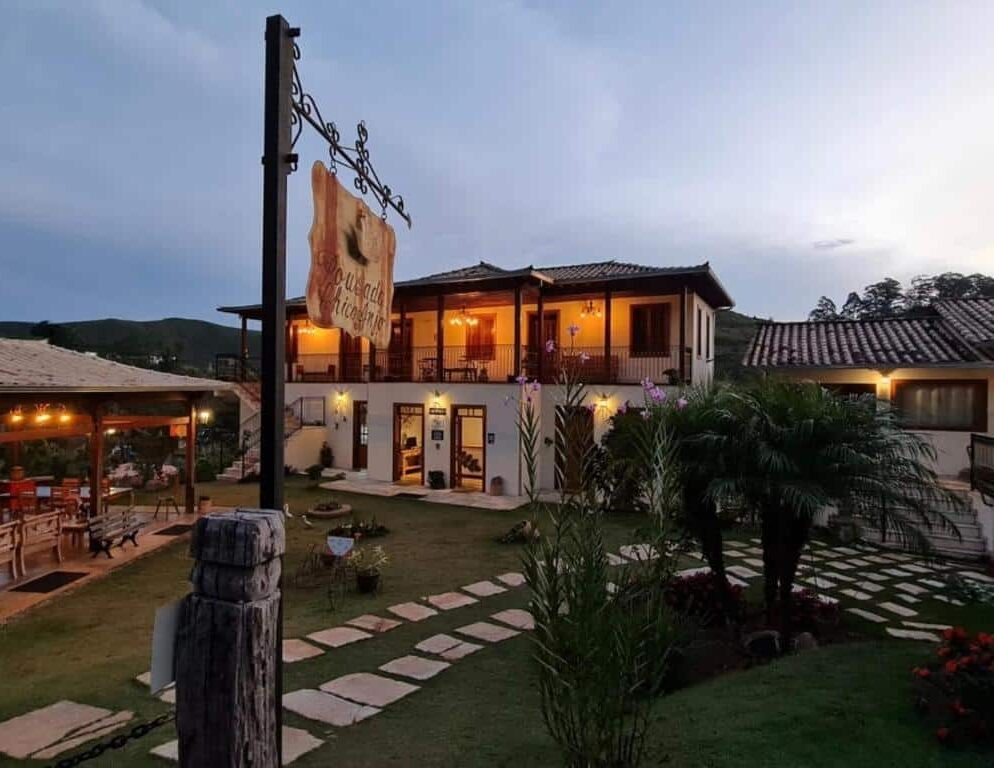 Um edifício rústico de dois andares da Pousada Chico Anjo com varandas e assentos ao ar livre, cercado por um jardim ao anoitecer.