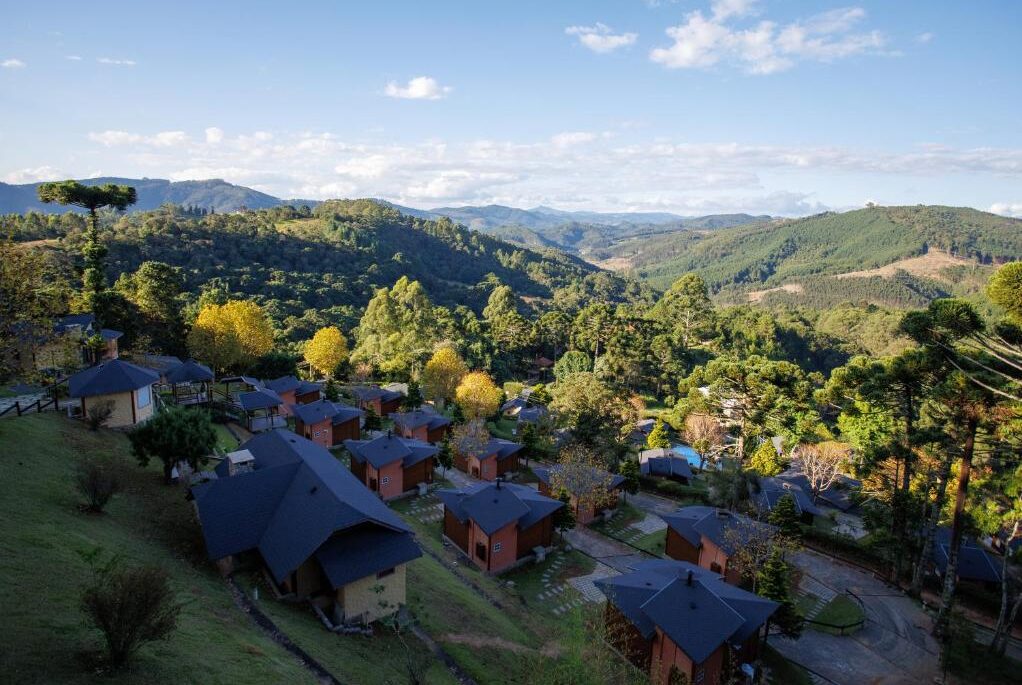As cabanas com telhados escuros estão aninhadas em uma encosta cercada por árvores e montanhas verdejantes sob um céu azul na Pousada Varanda das Colinas.