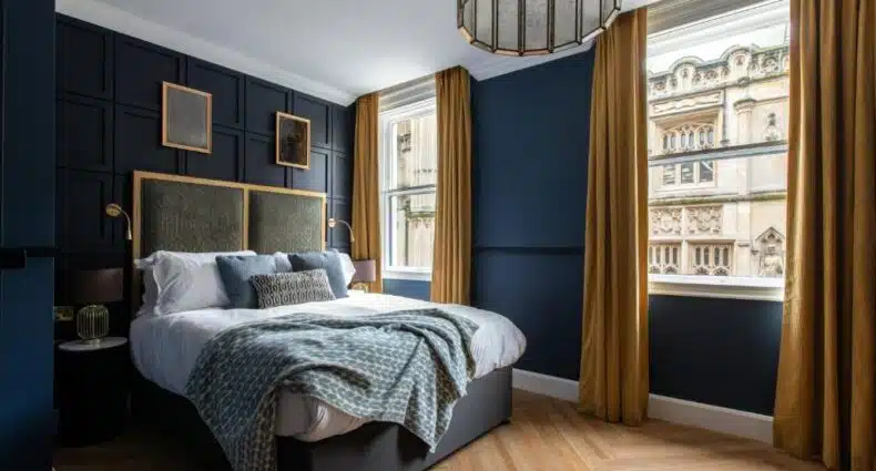 Um quarto do Hort's Townhouse com uma cama de casal, cortinas douradas, paredes azuis escuras, piso de madeira e duas grandes janelas voltadas para edifícios históricos. Imagem para ilustrar post sobre hotéis em Bristol.