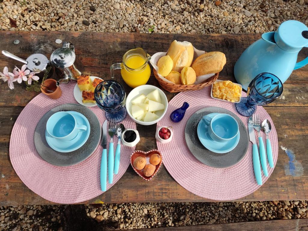 Uma mesa de madeira rústica posta para duas pessoas com pratos azuis, pão, suco e itens de café da manhã em jogos americanos cor-de-rosa.