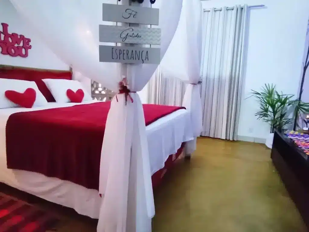 Um quarto com uma cama vermelha e branca, travesseiros em forma de coração, arte na parede, uma planta e um dossel transparente sobre a cama.