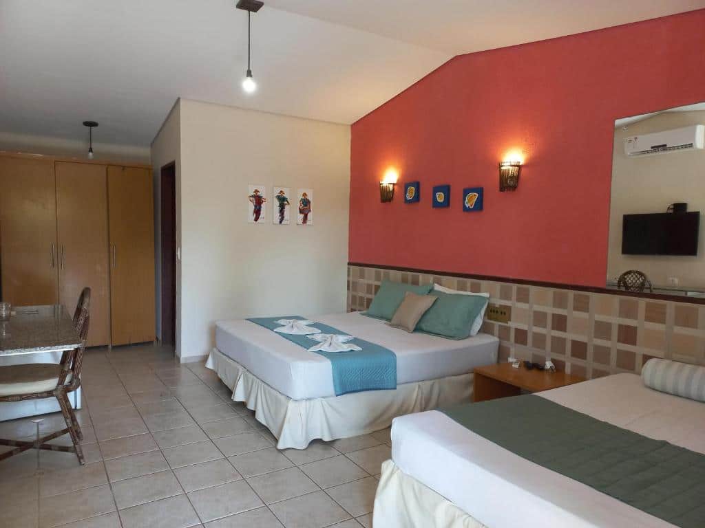 Hotel Enseada Maracajaú. Quarto de hotel com duas camas, uma parede com detalhes em vermelho, toalhas dobradas, arte na parede, uma mesa com cadeiras e um guarda-roupa.