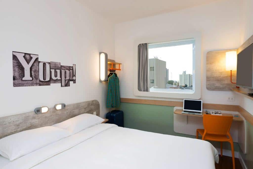 Um quarto do ibis budget Campinas Aquidaban com uma cama de casal, escrivaninha, cadeira laranja, cabideiro, janela e arte na parede que diz "Youpi!". Essa é uma das recomendações de onde ficar em Campinas.