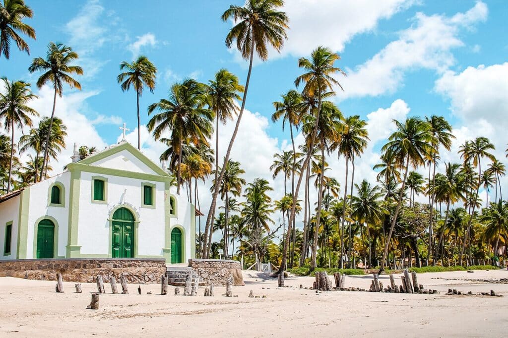 Uma igreja branca com portas verdes fica em uma praia de areia cercada por palmeiras altas sob um céu parcialmente nublado.