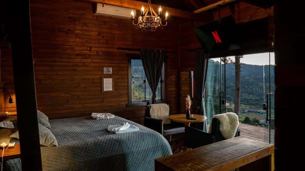 Um quarto aconchegante em cabine de madeira no Infinity Valley Cabanas de Altitude Urubici com cama, lustre, TV, duas cadeiras, uma pequena mesa e janelas grandes com vista para a natureza.