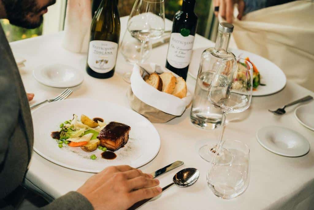 Uma pessoa se senta em uma mesa do Mastrojanni Relais com vinho, água, pão e uma refeição servida em um restaurante fino. Imagem para ilustrar post sobre hotéis românticos na Toscana.