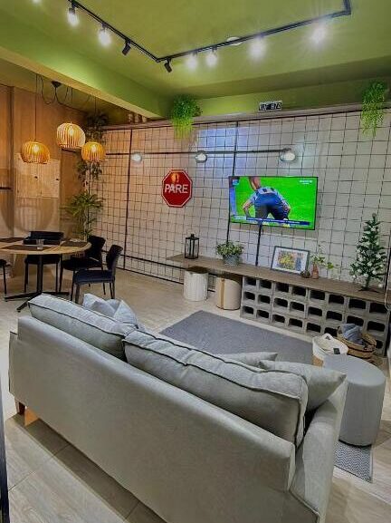 Sala de estar moderna do São Hostel com sofá de cor clara, TV em uma parede de azulejos, área de jantar e plantas; placa "PARE" na parede.