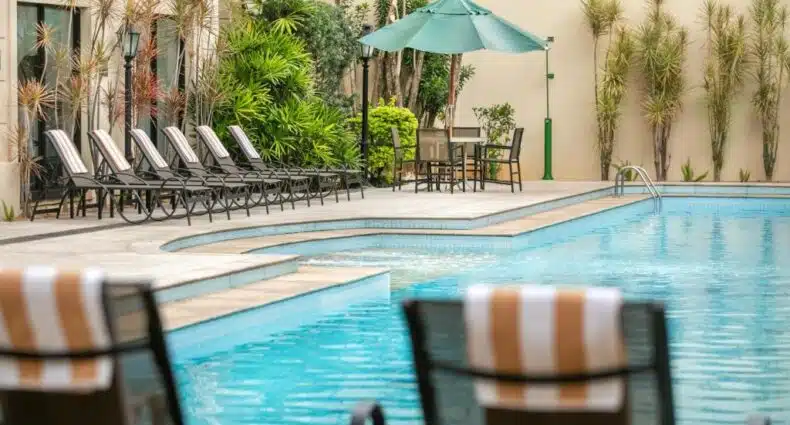 Área da piscina externa do Meliá Campinas com espreguiçadeiras, toalhas listradas, mesa com cadeiras, guarda-sol e plantas ao redor. Essa é uma das recomendações de onde ficar em Campinas.