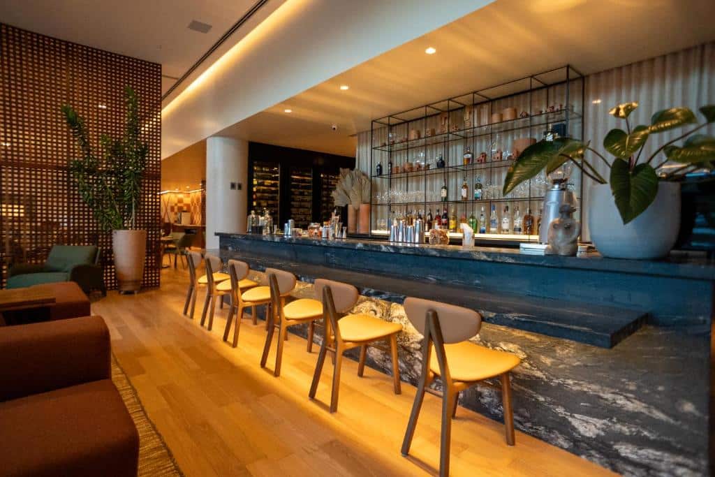 Bar no Monã Hotel com banquetas de madeira, balcão de pedra, prateleiras de garrafas e plantas em um lounge contemporâneo e bem iluminado. A foto faz parte do post de onde ficar em Teresina.