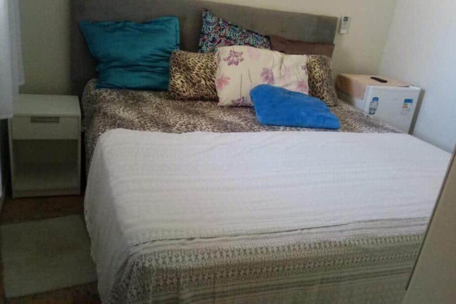 Paraíso de Maracajaú 1. Uma cama bem arrumada com vários travesseiros, uma toalha azul, uma pequena mesa de cabeceira e um frigobar ao lado.