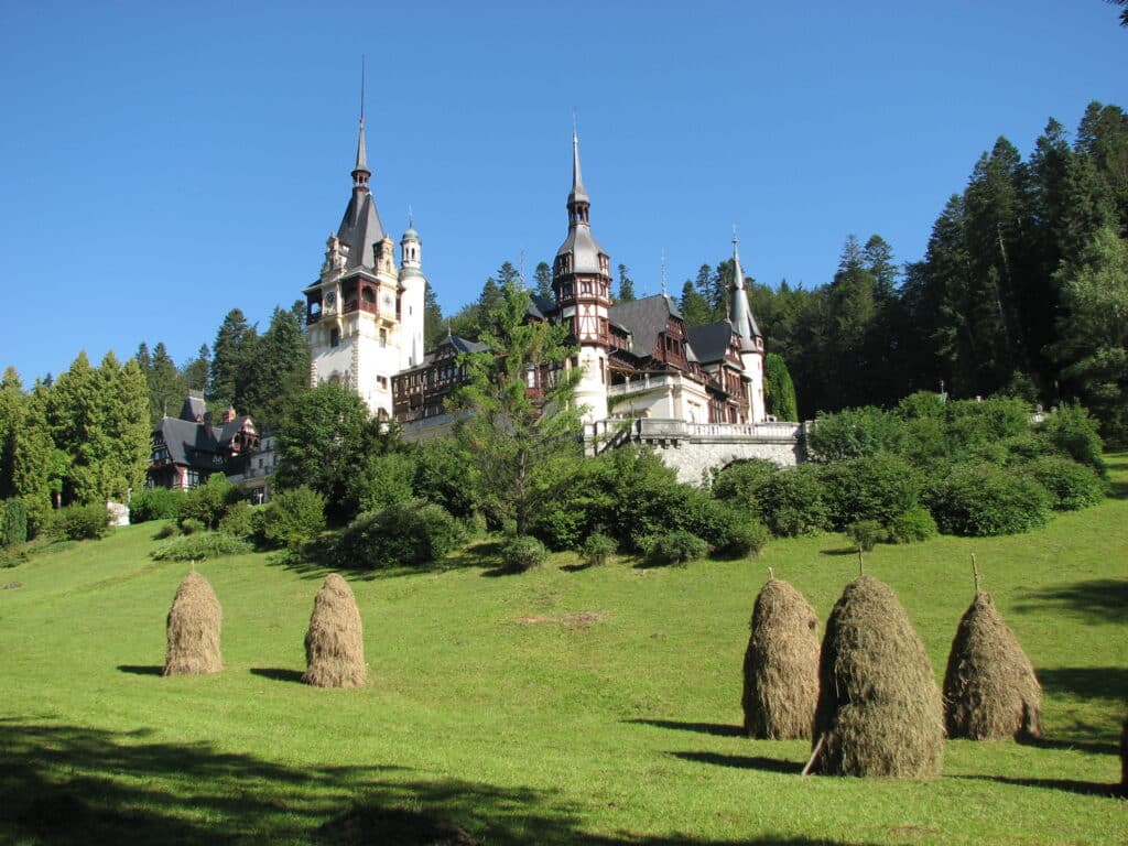 Peleș Castle com altas torres em uma colina verde, cercado por árvores, com palheiros no primeiro plano gramado sob um céu claro.