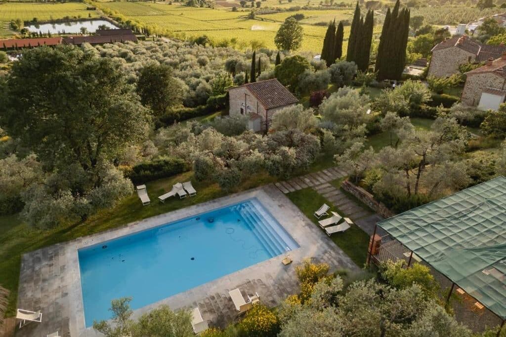 Uma piscina externa retangular do Il Palazzo é cercada por espreguiçadeiras, árvores e edifícios com uma paisagem rural ao fundo.