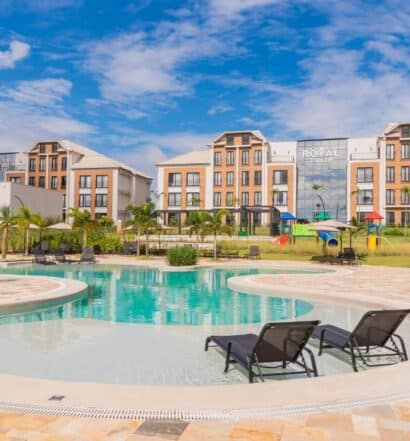 Duas espreguiçadeiras em uma piscina curva em frente a um moderno edifício de hotel com palmeiras e um playground, na propriedade do Royal Tulip Holambra, para representar onde ficar em Holambra.