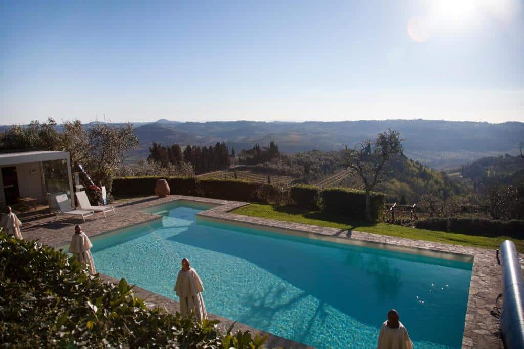 Piscina externa retangular da Villa I Barronci Resort & Spa com espreguiçadeiras, cercada por vegetação, com vista para as colinas sob um céu claro. Imagem para ilustrar post sobre hotéis românticos na Toscana.