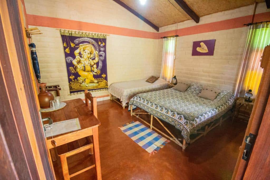 Um quarto aconchegante na Pousada Aruana com duas camas, uma pequena escrivaninha, tapetes coloridos e uma decoração de parede com uma divindade hindu.