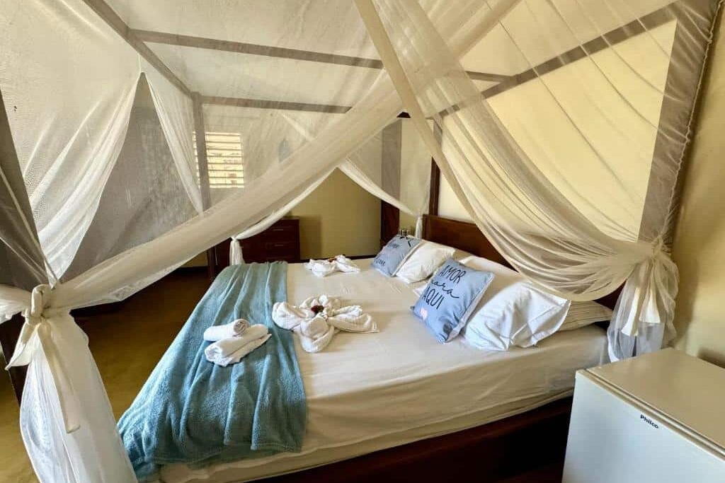Pousada Bibiku. Uma cama com mosquiteiro, toalhas dobradas, cobertor azul, travesseiros e uma pequena geladeira branca em um quarto.
