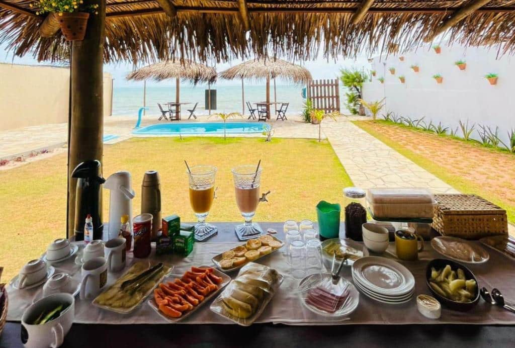 Buffet de café da manhã servido em uma mesa sob um telhado de palha, com piscina, pátio e oceano ao fundo. Foto da pousada bibiku