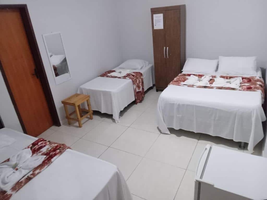 Um quarto da Pousada Canoa de Tolda simples com três camas, um guarda-roupa, um pequeno banco de madeira, um espelho e um frigobar branco.