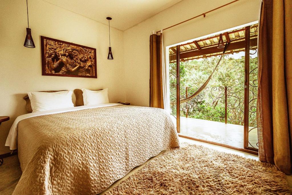 Um quarto na Pousada Casa de Shiva com uma cama grande, roupa de cama bege, arte na parede, um tapete e uma porta de vidro deslizante que se abre para uma varanda com uma rede. A foto é parte do post de onde ficar na Chapada dos Veadeiros.