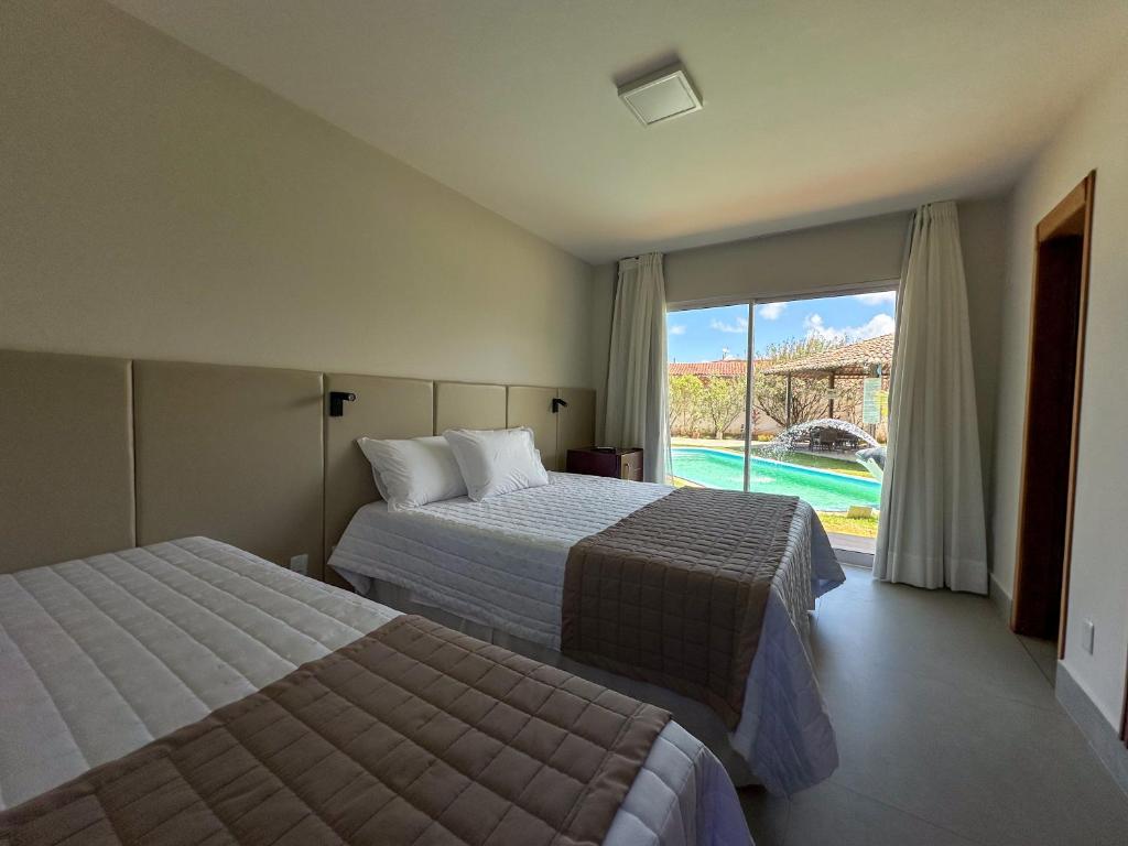Pousadas Corais. Um quarto de hotel com duas camas, roupa de cama branca, detalhes em bege e uma janela com vista para a piscina e a área externa.