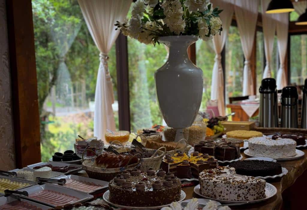 Uma mesa da Pousada Encanto do Avencal cheia de bolos, doces e frios, com um vaso alto de flores brancas no centro. Essa é uma das dicas de onde ficar em Urubici.