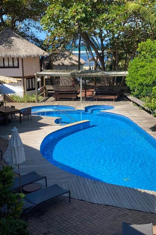 Piscina externa da Pousada Farol das Tartarugas com formato curvo, deck de madeira, espreguiçadeiras, guarda-sóis e árvores, com o oceano visível ao fundo.