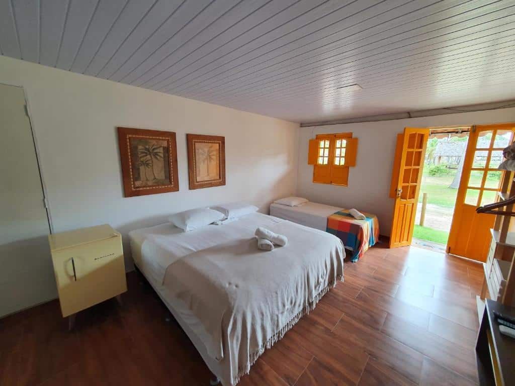 Um quarto na Pousada Igrejinha dos Carneiros com uma cama de casal, uma cama de solteiro, um pequeno frigobar, quadros de arte e uma porta aberta para uma área externa gramada. Essa é uma das indicações de onde ficar na Praia dos Carneiros.