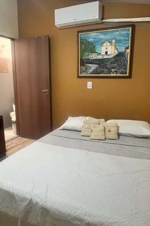 Uma cama bem arrumada em um quarto com paredes cor de bronze, um quadro, ar-condicionado e uma porta aberta que dá para um banheiro.