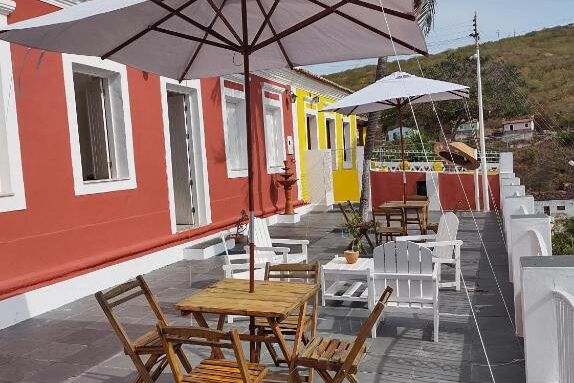Pousada Maria Bonita. Pátio externo com mesas de madeira, cadeiras, guarda-chuvas brancos e um prédio colorido vermelho e amarelo ao fundo.