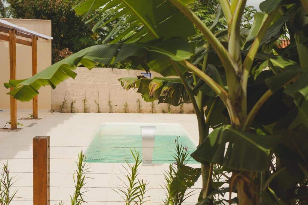 Uma pequena piscina externa na Pousada Paraiso com um recurso de água é cercada por um pátio de azulejos, plantas e uma parede bege sob um céu claro.