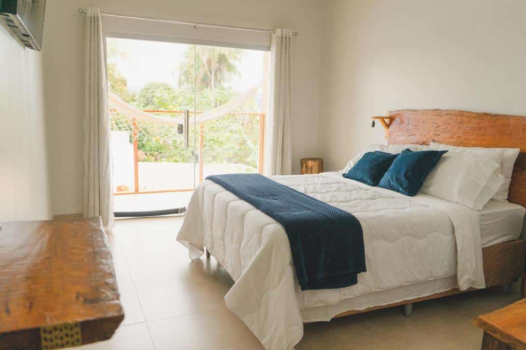 Uma cama bem arrumada na Pousada Paraiso com detalhes em azul em um quarto bem iluminado; uma rede pode ser vista do lado de fora através de portas de vidro. A foto é parte do post de onde ficar na Chapada dos Veadeiros.