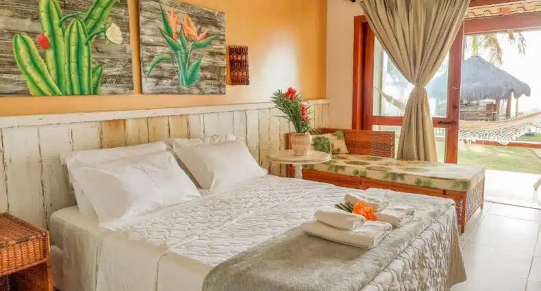 Uma cama bem arrumada em um quarto bem iluminado com decoração tropical, móveis de vime e vista para uma rede do lado de fora.