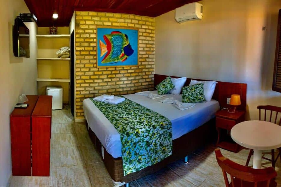 Pousada Ponta dos Anéis. Um quarto de hotel com uma cama de casal, roupa de cama verde, uma parede de tijolos, uma pequena mesa com duas cadeiras e um frigobar.