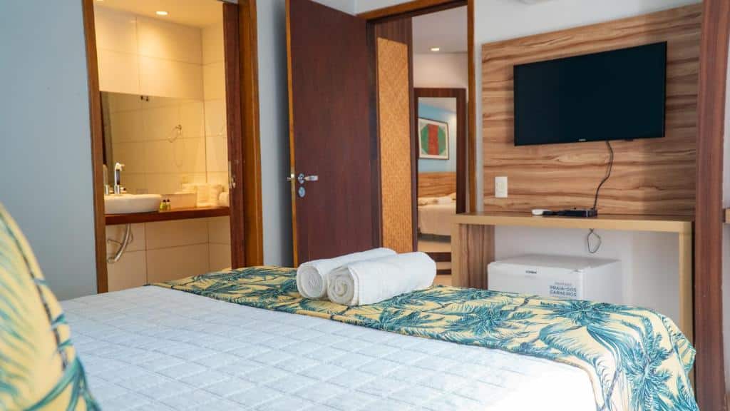 Um quarto da Pousada Praia dos Carneiros com uma cama, toalhas enroladas, uma TV na parede, um frigobar e uma porta aberta que mostra um banheiro. Essa é uma das indicações de onde ficar na Praia dos Carneiros.